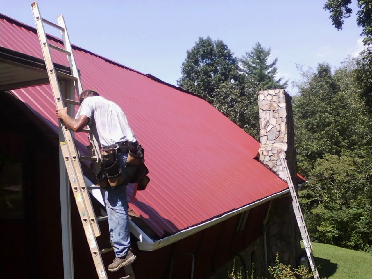 Roofer on a ladder beside a red metal roof and stone chimney — Soffit & Fascia Repair by Roofing Brothers in Ransom